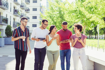 Obraz premium Cheerful friends walking together with gadgets. Men and women using smartphones and tablet, discussing news and laughing. Mobile technology concept