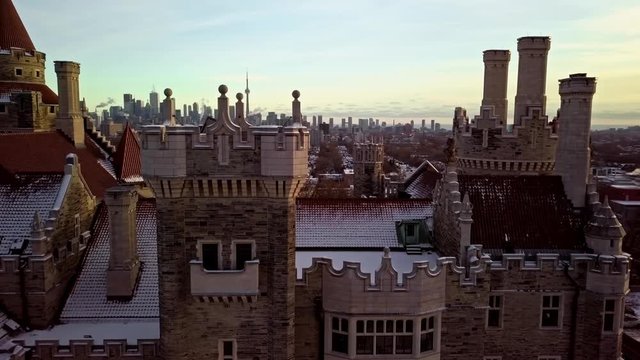 Contrast between Old Castle and Modern City at Sunset, Drone Parallax. Flying past the Turrets and Red Tile Roof of a Snowy Mansion Home with the Downtown Toronto Skyline behind in Winter Golden Hour