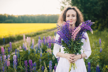 Beautiful woman in a white dress holds a large bouquet of lupins in the field.