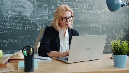 Mature woman is using laptop in class working online typing sitting at desk alone concentrated on work. Modern technology and communication concept. - Powered by Adobe