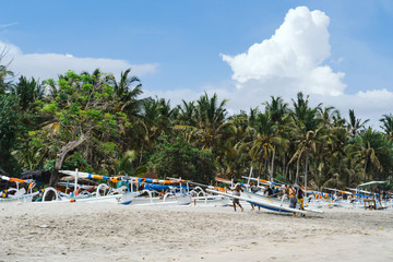 Fototapeta na wymiar Fishermen carry a boat on a virgin beach in Bali. Fishing boats on white sand. Coconut trees and blue sky with clouds on the background. Clear sunny day
