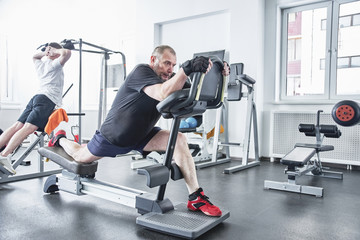Obraz na płótnie Canvas Adult gray-haired man trains on fitness equipment in the gym, pumps legs and arms muscles, loses weight. Concept of healthy lifestyle in old age