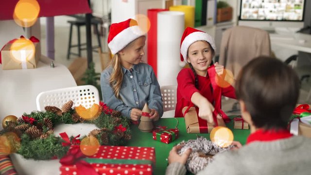 Two little girls in Santa hats talking and decorating Christmas presents together with female designer during craft workshop in studio; garland lights flickering in the foreground