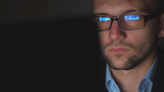 Male face of manager in eyeglasses with reflection of screen lights working on laptop late at night. Young businessman looking at monitor using modern computer in office. Close up Slow motion