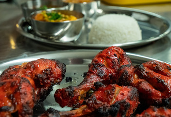 Close-up shot of a plate of cooked tandoori chicken in the foreground with rice and other Indian food in the background.