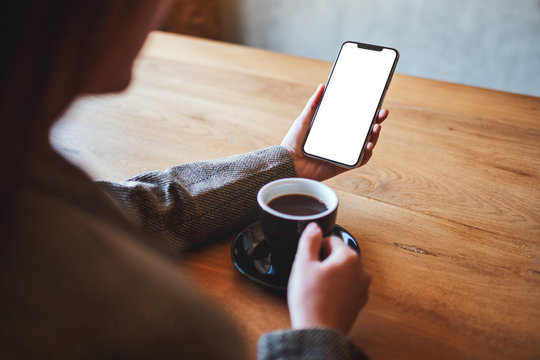 Mockup image of a woman holding black mobile phone with blank screen while drinking coffee on wooden table