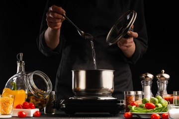 Cooking soup. A cook prepares soup in a saucepan against the background of ingredients. Delicious and healthy food. Black background. Salt, freezing in motion. Culinary and gastronomy.