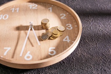 Time is money concept. Wooden clock with coins on dark textured background, sunny light. Saving money concept, banking and business idea. Copy space.