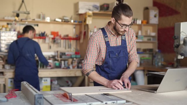 Lockdown of young Caucasian carpenter with beard and glasses drawing lines on plank and using laptop for checking, while man on background doing something with his hands