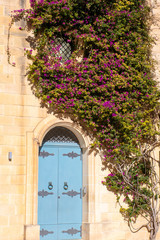 Fototapeta na wymiar Blue door and purple flowers covering stone wall in Mdina Malta