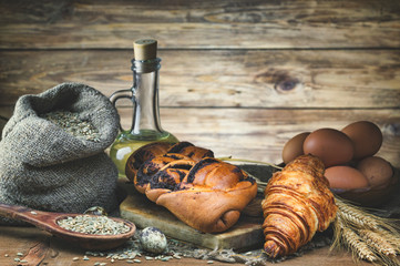Sweet bread, croissant with poppy seeds, wheat ear, egg, and grain in a bag lie on a wooden rustic table against a  wooden background. Low key. Toned. Focus concept