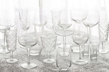 Several rows clear of glasses for wine, champagne, water and other drink on grey concrete table at backlighting. Abstract tableware background
