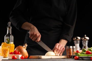 Chef cook slices onions in the kitchen on a dark background with vegetables, close-up