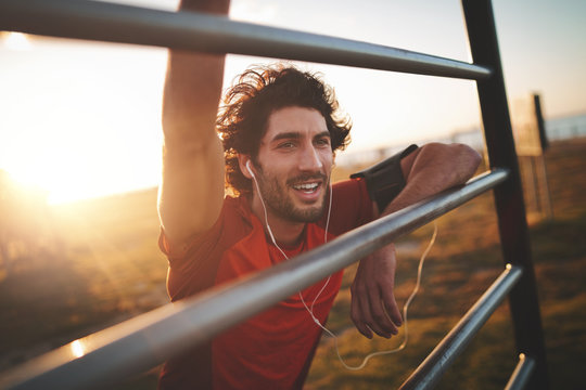 Portrait of a cheerful young handsome male runner with earphones in his ears leaning on railing after exercising in the park on a sunny day