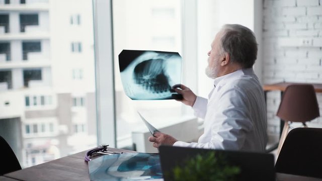 Experienced adult doctor with thick white beard siting at desk and examining X-ray scan against large window in modern office room of the hospital. Shooting in slow motion.