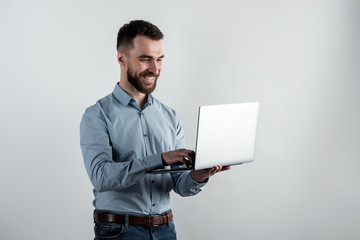 Cheerful guy in blue shirt stands holding an open  laptop against a white studio background, looking at the screen © speed300