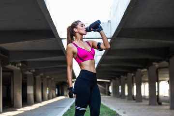 Beautiful female runner standing outdoors drinking water from her bottle. Fitness woman taking a break after running workout.Athletic young woman in sports dress doing fitness exercise.