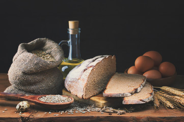 Rye bread with grain in a wooden spoon lies on a wooden rustic table on a dark background. The bread lies on a coarse sackcloth with wheat ears next to a bag of grain and quail and chicken eggs.