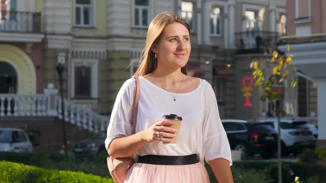 4k slow motion video of happy smiling woman with long hair drinking coffee to go during office break while walking on city street