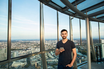 Obraz premium Young man in tshirt stands on the observation deck, holding cellphone and typing text message. On the background panorama of Frankfurt city. Germany
