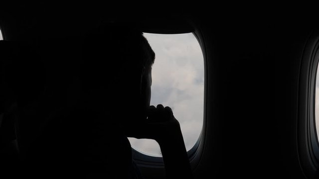 Passenger man sit against window and watch out to clouds from above, travelling by regular airline. Silhouetted shot, thoughtful guy hold hand at chin, lost in dreams