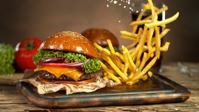 Super Slow Motion Shot of Fast Food Concept. Falling French Fries on Wooden Cutting Board next to the Fresh Hamburger at 1000fps.