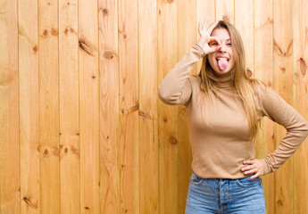young blonde woman smiling happily with funny face, joking and looking through peephole, spying on...