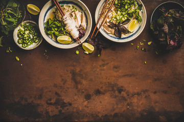 Asian cuisine lunch. Flat-lay of Vietnamese rice noodle chicken soup Pho Ga with fresh cilantro, soy sprouts, greens, lime in bowls with chopsticks over rusty dark background, top view, copy space