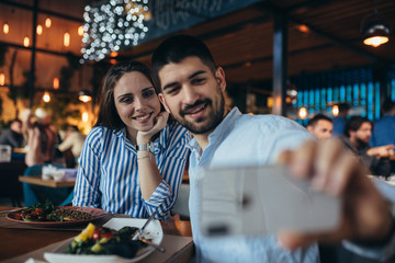 happy young couple taking picture with mobile phone while having lunch in restaurant