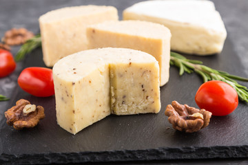 Cheddar cheese and various types of cheese with rosemary and tomatoes on black slate board on a black concrete background. Side view, selective focus.