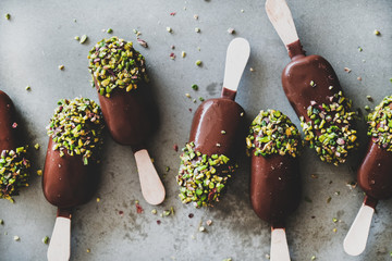 Ice cream popsicle pattern. Flat-lay of chocolate glazed ice cream pops with pistachio icing over grey concrete background, top view, close-up. Summer seasonal cold sweet healthy vegan dessert