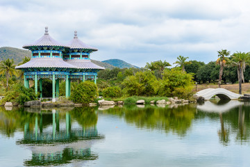 Fototapeta na wymiar Green lake surrounded by gazebos, temples, mountains, flowers, palm grove and stone fence on the territory of Buddhist center Nanshan.