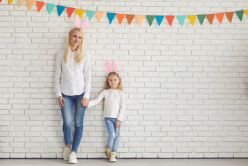 Obraz na płótnie Canvas Happy easter. Mother and daughter with rabbit ears smiling against a white brick wall.