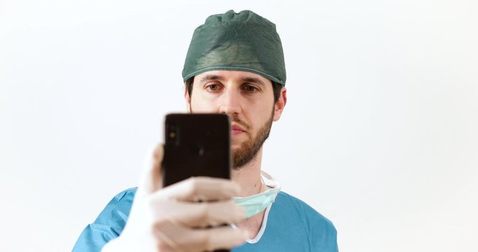 Giovane medico chirurgo con barba lunga con lo smartphone si fa i video selfie con il telefonino, sfondo bianco.