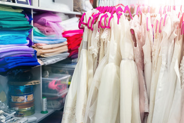 Rack with many beautiful holiday dresses for girls on hangers and multicolored fabric stack at children fashion sewing studio and showroom indoor. Female dress rental salon maker photo session event