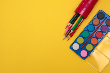 A palette of paints for drawing and color pencils in a pencil case on a yellow background, space for text. Flat lay.