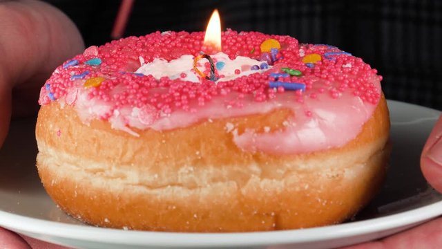 Hands holding a cake made out of a donut with candy and icing with a birthday candle lighted on top. Pink background.