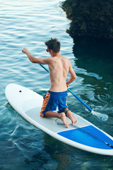 Obraz premium Stand Up Paddling in the sea. SUP. Guy Training in the morning on Paddle Board near the rocks.