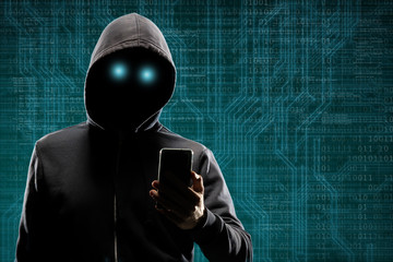 Dangerous hacker with a smartphone gadget over digital background with binary code. Obscured dark face in mask and hood. Data thief, internet attack, darknet fraud, virtual reality and cyber security.