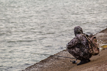 adult in camouflage fishing on a river in a city. A real fisherman is fishing on a cloudy day. professional fisherman throws fishing rod