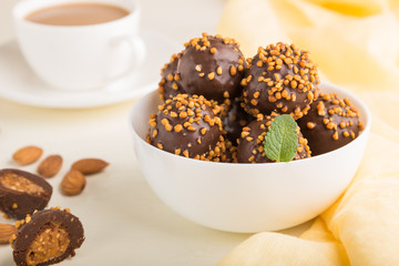 Chocolate caramel ball candies with almonds and a cup of coffee on a white wooden background. Side view, selective focus.