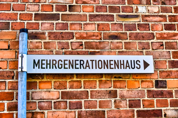 Street sign that points to a multi-generational house in which old and young people live together. The text Mehrgenerationenhaus is German for multi-generational house.