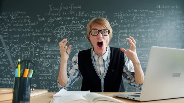 Portrait of excited guy laughing looking at camera sitting at table in university class, chalkboard with formulas is visible in the background. People and emotions concept.