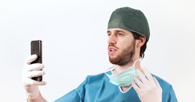 Medico chirurgo uomo con barba lunga e mascherina da sala operatoria fa una videochiamata con il suo smartphone. Sfondo bianco.