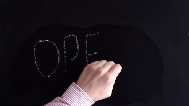 A hand with chalk writes the word closed on the Board, then wipes it with a wet rag and writes it open. The word is closed on the Board written in chalk, wipe with a damp cloth and write open