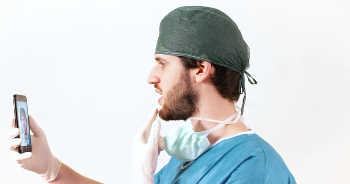 Giovane medico chirurgo con barba lunga con lo smartphone si fa i video selfie con il telefonino, sfondo bianco.