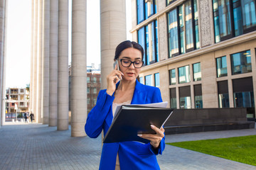 Confident woman skilled banker reading financial information from paper documents and talking via cell telephone while standing outdoors near contemporary enterprise building