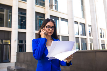 Woman in glasses skilled CEO reading information from paper documents and talking via mobile phone while standing outside business centre during work break