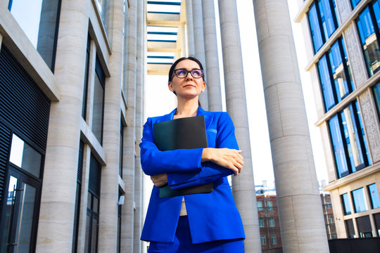 Bottom view of a serious pensive business woman in glasses and suit holding folder with paper documents and thrustful looking away while standing outdoors near modern enterprise building
