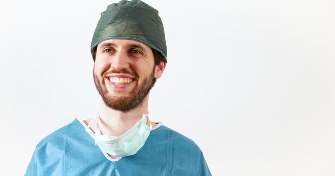Medico chirurgo uomo con mascherina abbassata e barba guarda preoccupato intorno a se e poi sorride di fronte la telecamera. Sfondo bianco.